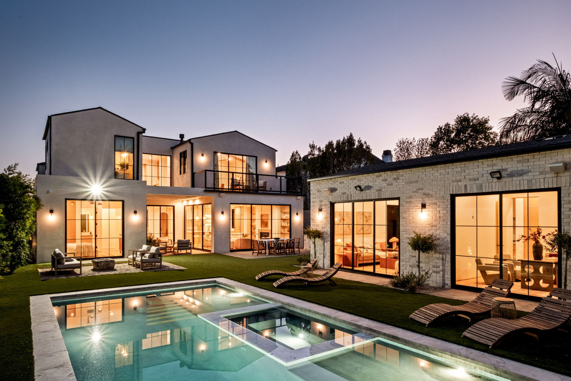 Modern Spanish-style residence at dusk with pool, lit steel windows and brick guest house